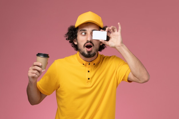 Mensajero masculino de vista frontal en uniforme amarillo y capa sosteniendo la taza de café de entrega y la tarjeta en la pared rosa