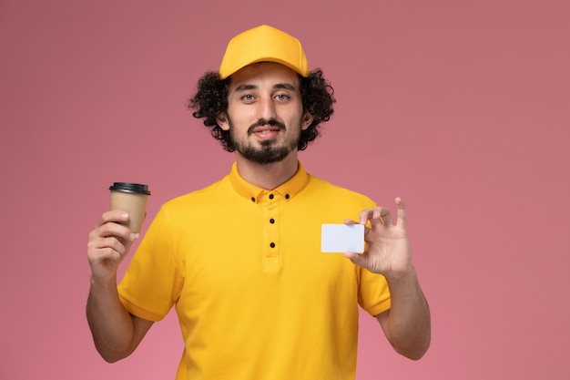 Mensajero masculino de vista frontal en uniforme amarillo y capa sosteniendo la taza de café de entrega y la tarjeta en la pared rosa
