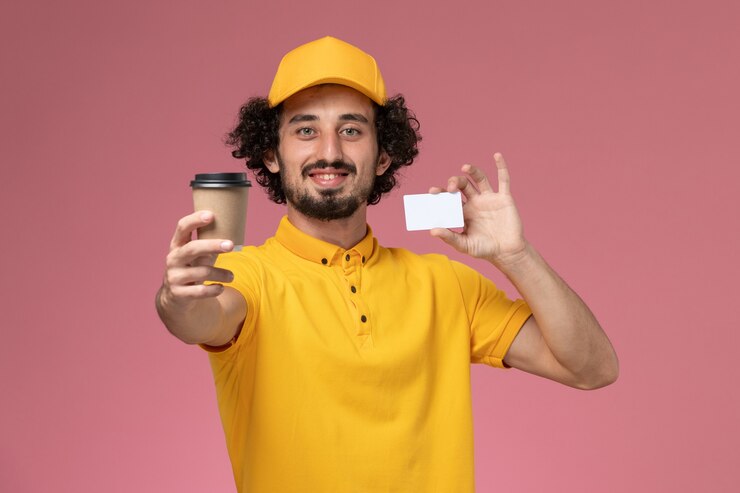 Foto gratuito mensajero masculino de vista frontal en uniforme amarillo y capa sosteniendo la taza de café de entrega y la tarjeta en la pared rosa