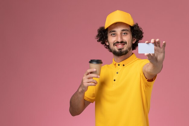 Mensajero masculino de vista frontal en uniforme amarillo y capa sosteniendo la taza de café de entrega y la tarjeta en la pared rosa