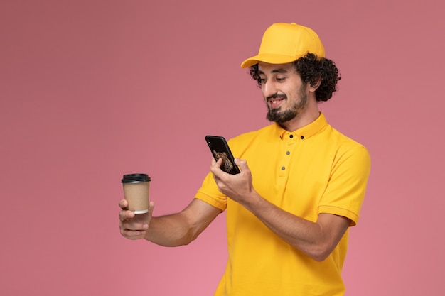 Foto gratuita mensajero masculino de vista frontal en uniforme amarillo y capa sosteniendo la taza de café de entrega tomando una foto en la pared rosa