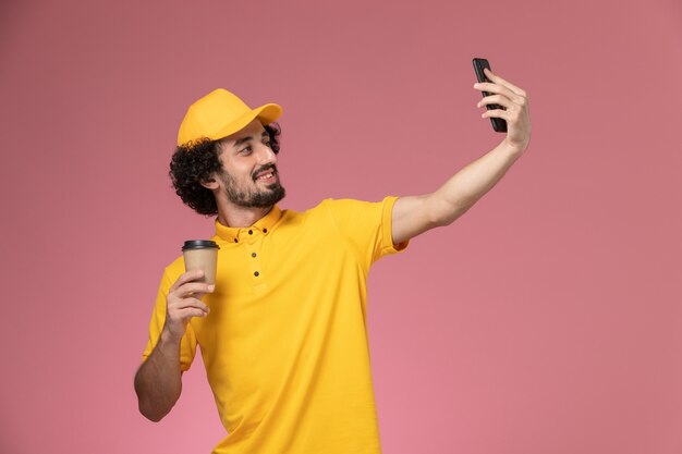 Mensajero masculino de vista frontal en uniforme amarillo y capa sosteniendo la taza de café de entrega tomando selfie en la pared rosa