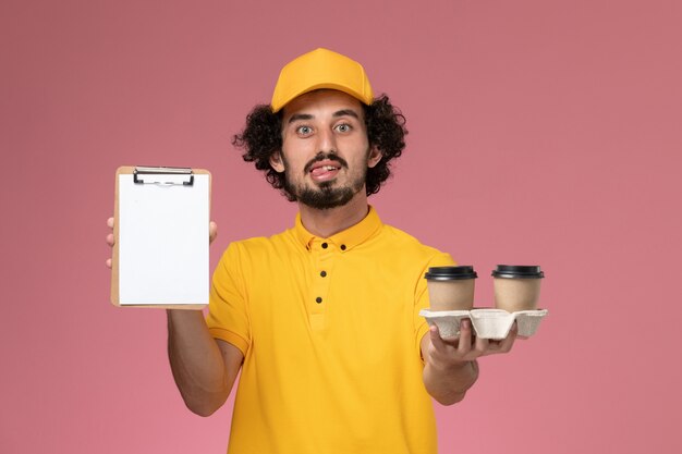 Mensajero masculino de vista frontal en uniforme amarillo y capa sosteniendo tazas de café de entrega marrón y bloc de notas en la pared rosa