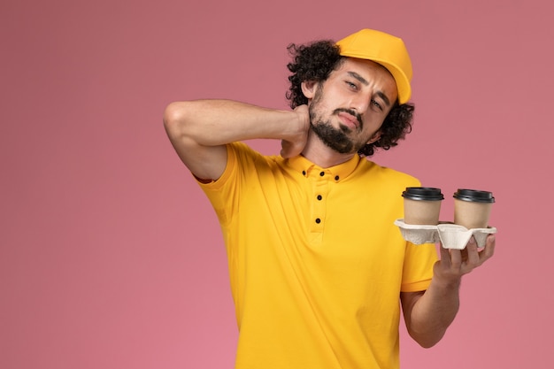 Mensajero masculino de vista frontal en uniforme amarillo y capa sosteniendo tazas de café de entrega marrón en la pared rosa