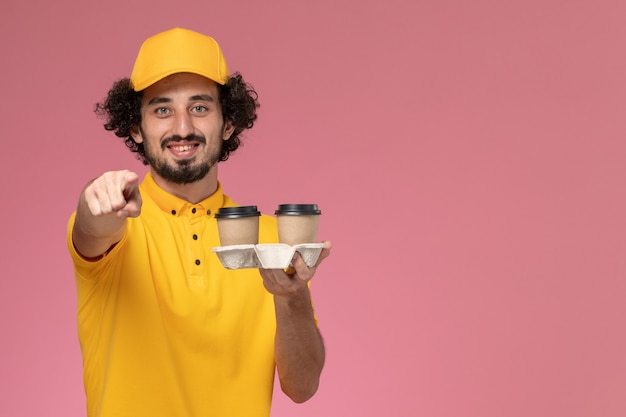 Mensajero masculino de vista frontal en uniforme amarillo y capa sosteniendo tazas de café de entrega marrón con una sonrisa en la pared rosa