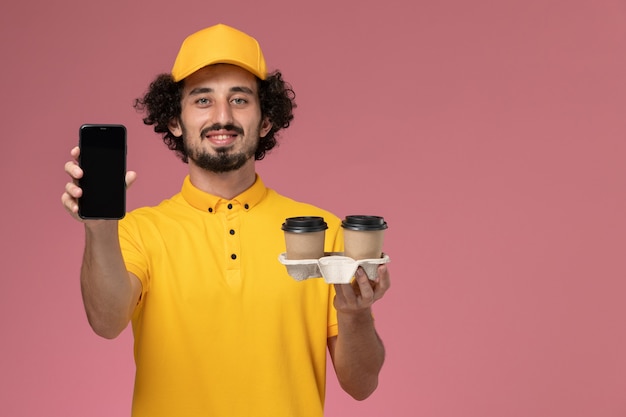 Mensajero masculino de vista frontal en uniforme amarillo y capa sosteniendo tazas de café de entrega marrón y teléfono en la pared rosa