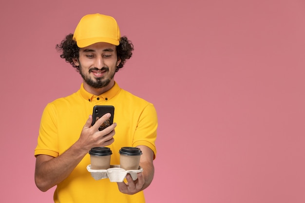 Mensajero masculino de vista frontal en uniforme amarillo y capa sosteniendo tazas de café de entrega marrón y tomando fotos de ellos en la pared rosa