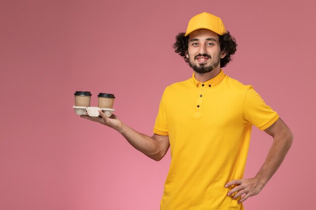 Mensajero masculino de vista frontal en uniforme amarillo y capa sosteniendo tazas de café de entrega en la pared rosa