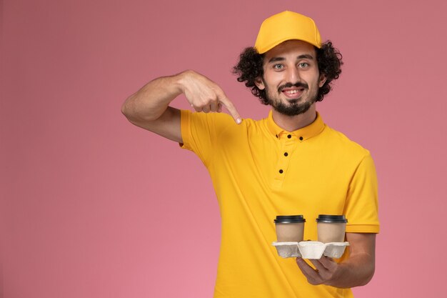 Mensajero masculino de vista frontal en uniforme amarillo y capa sosteniendo tazas de café de entrega en la pared rosa