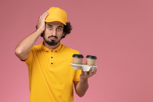 Mensajero masculino de vista frontal en uniforme amarillo y capa sosteniendo tazas de café de entrega en la pared rosa