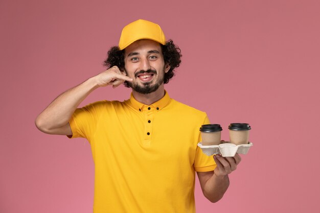 Mensajero masculino de vista frontal en uniforme amarillo y capa sosteniendo tazas de café de entrega en la pared rosa