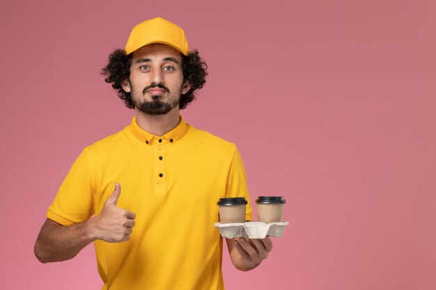 Mensajero masculino de vista frontal en uniforme amarillo y capa sosteniendo tazas de café de entrega en la pared rosa