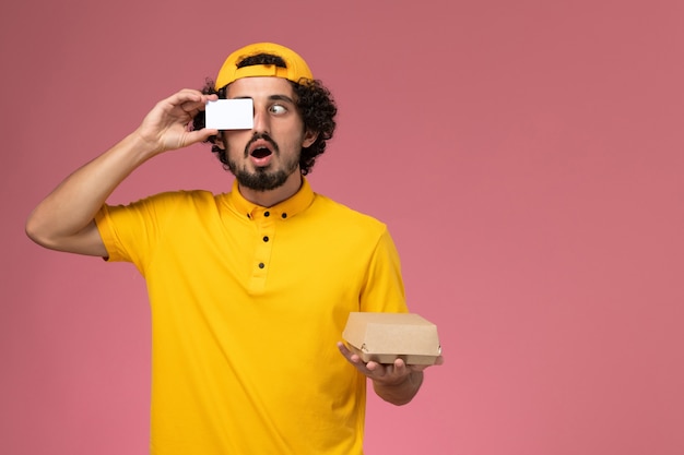 Mensajero masculino de vista frontal en uniforme amarillo y capa con tarjeta y pequeño paquete de comida de entrega en sus manos sobre el fondo rosa claro.