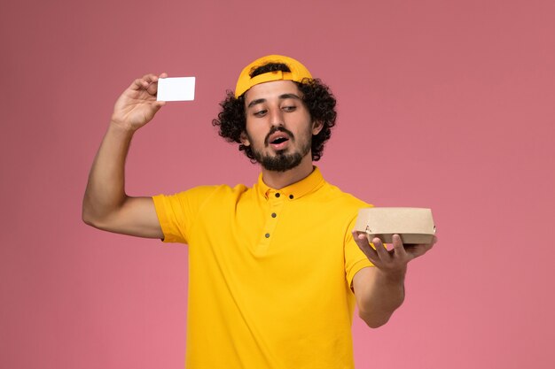 Mensajero masculino de vista frontal en uniforme amarillo y capa con tarjeta y pequeño paquete de comida de entrega en sus manos sobre el fondo rosa claro.