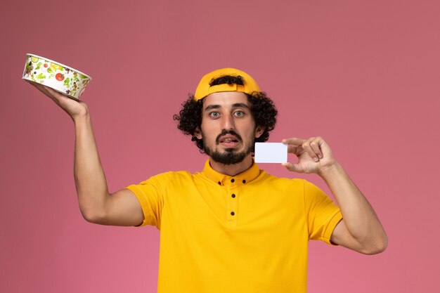 Mensajero masculino de vista frontal en uniforme amarillo y capa con tarjeta de tazón de entrega redonda en sus manos sobre el fondo rosa.