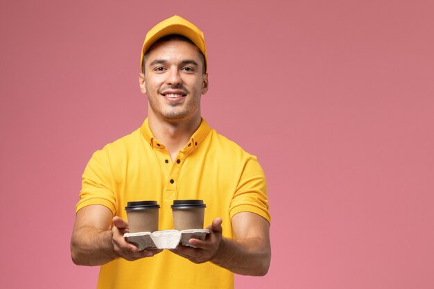 Mensajero masculino de vista frontal en uniforme amarillo entregando tazas de café de entrega en el fondo rosa claro