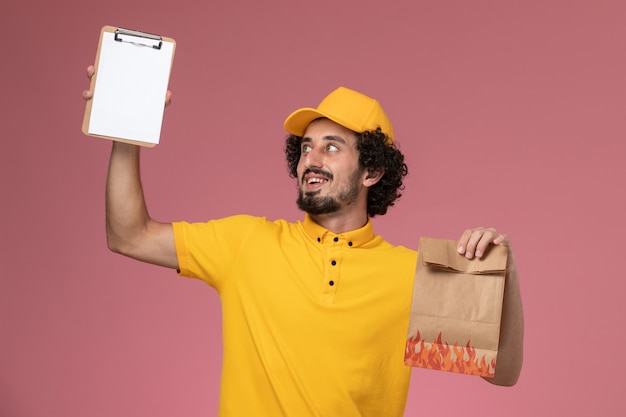 Mensajero masculino de vista frontal en uniforme amarillo con paquete de alimentos y bloc de notas en la pared rosa