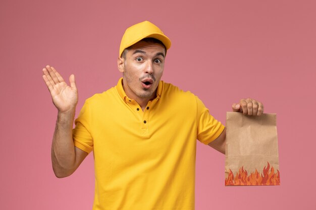 Mensajero masculino de vista frontal en uniforme amarillo con paquete de alimentos con expresión de sorpresa en el fondo rosa