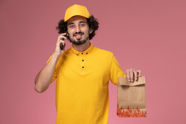 Mensajero masculino de vista frontal en uniforme amarillo con paquete de alimentos hablando por teléfono en la pared rosa