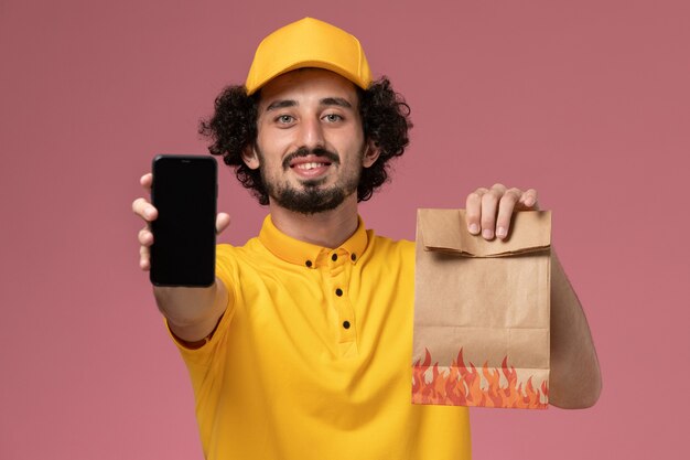 Mensajero masculino de vista frontal en uniforme amarillo con paquete de alimentos y teléfono inteligente en la pared rosa