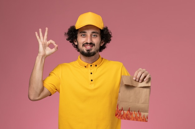 Mensajero masculino de vista frontal en uniforme amarillo con paquete de comida de papel en la pared rosa claro