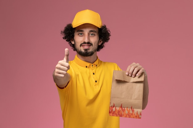 Mensajero masculino de vista frontal en uniforme amarillo con paquete de comida de papel posando en la pared de color rosa claro