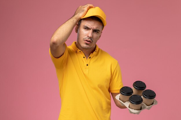 Mensajero masculino de vista frontal en uniforme amarillo pensando y sosteniendo tazas de café de entrega en el escritorio rosa