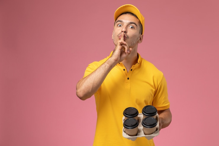Foto gratuito mensajero masculino de vista frontal en uniforme amarillo que muestra el signo de silencio y que sostiene las tazas de café de entrega en el escritorio rosa