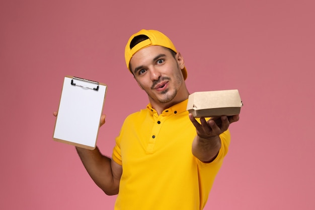 Foto gratuita mensajero masculino de la vista frontal en uniforme amarillo que sostiene el bloc de notas y el pequeño paquete de comida en el fondo rosa.