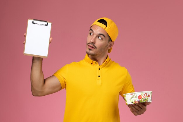 Mensajero masculino de la vista frontal en uniforme amarillo que sostiene el cuenco de la entrega junto con el bloc de notas en el fondo rosado.