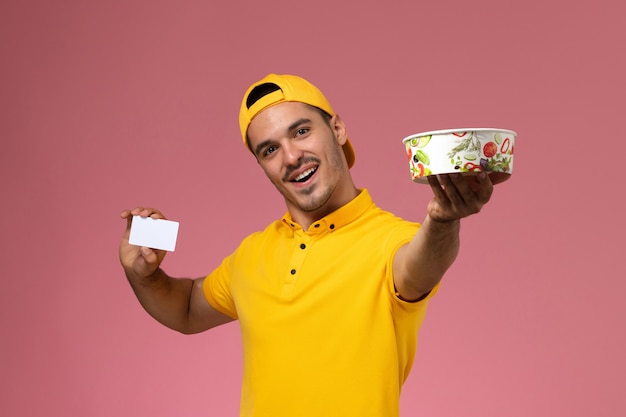 Mensajero masculino de la vista frontal en uniforme amarillo que sostiene el cuenco de la entrega con la tarjeta en fondo rosa claro.