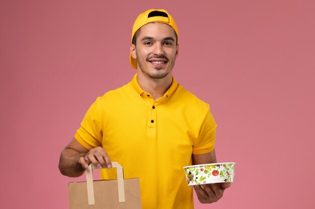 Mensajero masculino de la vista frontal en uniforme amarillo que sostiene el cuenco del paquete de alimentos de entrega sobre fondo rosa.