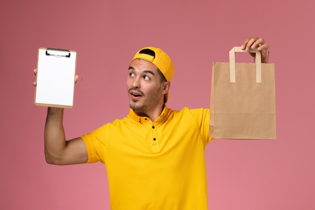 Mensajero masculino de la vista frontal en uniforme amarillo que sostiene el pequeño paquete de comida de la libreta y de la entrega en el escritorio rosado.