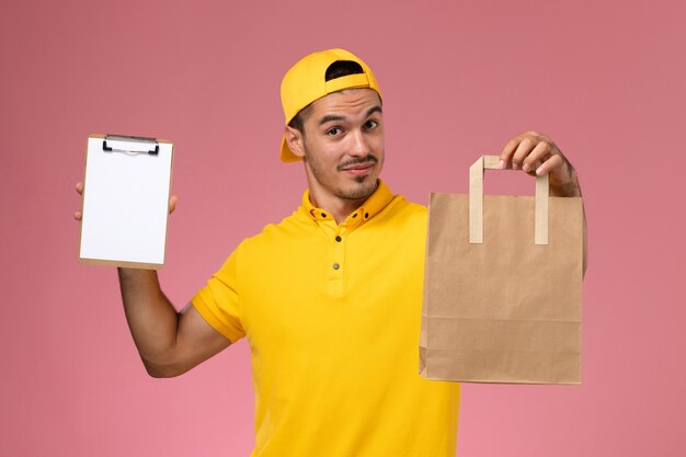 Mensajero masculino de la vista frontal en uniforme amarillo que sostiene el pequeño paquete de comida de la libreta y de la entrega en el fondo rosado.