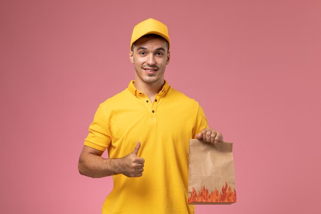 Mensajero masculino de vista frontal en uniforme amarillo sonriendo y sosteniendo el paquete de alimentos en el escritorio rosa