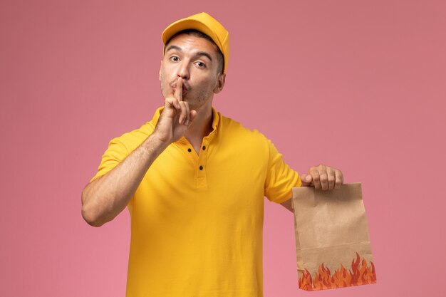 Mensajero masculino de vista frontal en uniforme amarillo sonriendo y sosteniendo el paquete de alimentos en el fondo rosa
