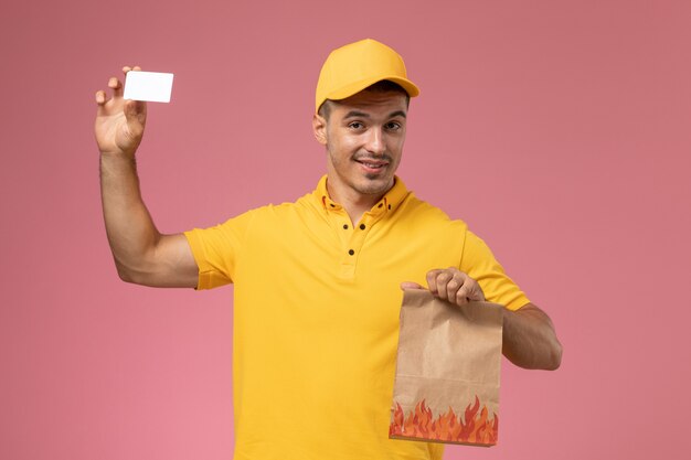 Mensajero masculino de vista frontal en uniforme amarillo sonriendo sosteniendo una tarjeta blanca y paquete de alimentos en el escritorio rosa