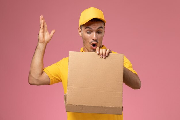 Mensajero masculino de vista frontal en uniforme amarillo sosteniendo y abriendo la caja de entrega de alimentos en el fondo rosa claro