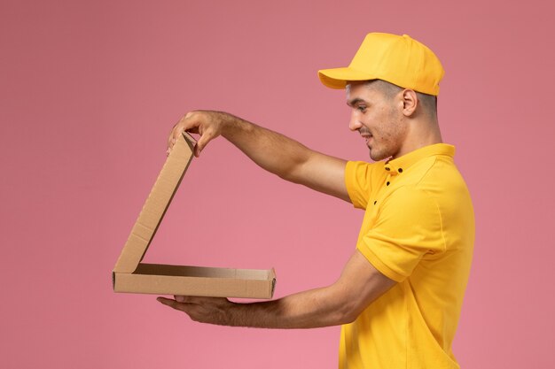 Mensajero masculino de vista frontal en uniforme amarillo sosteniendo y abriendo la caja de entrega de alimentos sobre fondo rosa