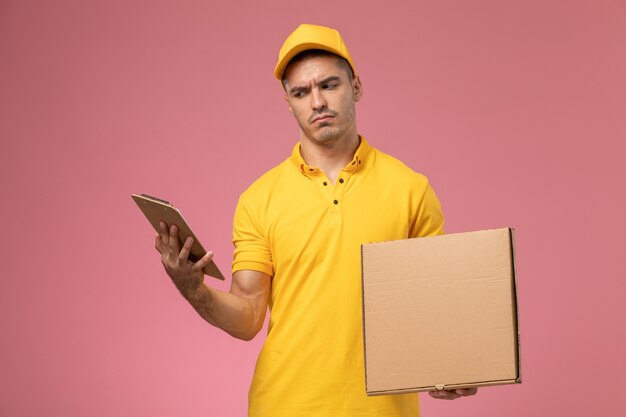 Mensajero masculino de vista frontal en uniforme amarillo sosteniendo el bloc de notas de lectura y la caja de entrega de alimentos en el fondo rosa