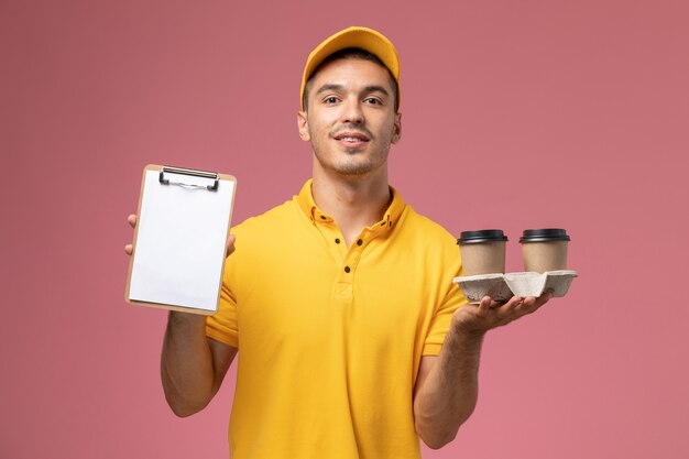 Mensajero masculino vista frontal en uniforme amarillo sosteniendo el bloc de notas y tazas de café de entrega sobre fondo rosa