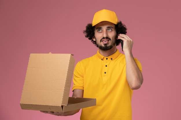 Mensajero masculino de vista frontal en uniforme amarillo sosteniendo la caja de entrega de alimentos y hablando por teléfono en la pared rosa claro