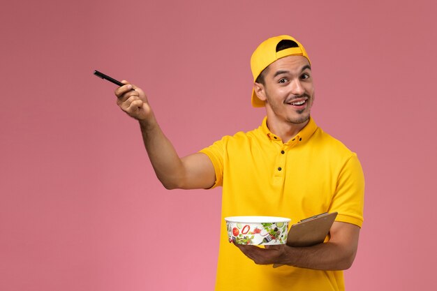 Mensajero masculino de vista frontal en uniforme amarillo sosteniendo el cuenco de entrega junto con el bloc de notas escribiendo notas sobre el fondo rosa.