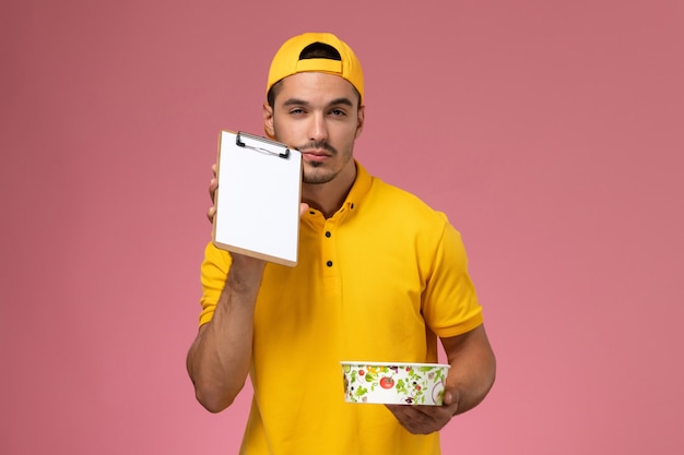 Mensajero masculino de vista frontal en uniforme amarillo sosteniendo el cuenco de entrega junto con el bloc de notas sobre fondo rosa claro.