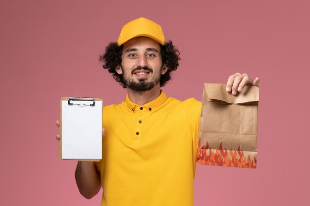 Mensajero masculino de vista frontal en uniforme amarillo sosteniendo el paquete de alimentos y el bloc de notas en la pared rosa claro
