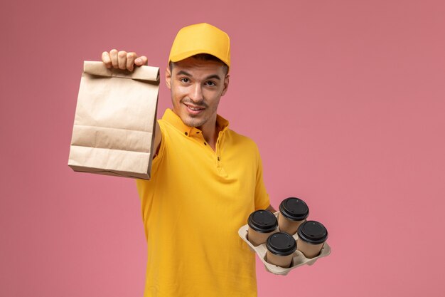 Mensajero masculino de vista frontal en uniforme amarillo sosteniendo el paquete de alimentos y tazas de café de entrega en el fondo de color rosa