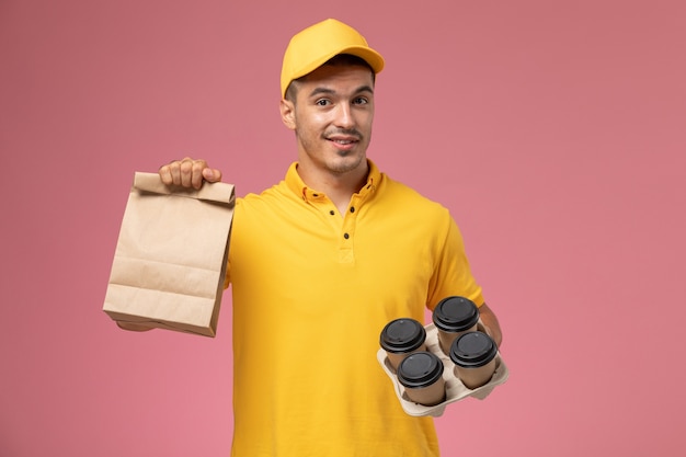 Mensajero masculino de vista frontal en uniforme amarillo sosteniendo el paquete de alimentos y tazas de café de entrega en el fondo de color rosa