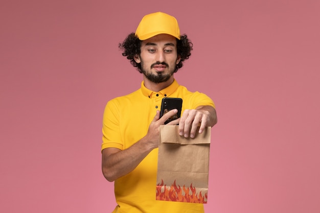 Mensajero masculino de vista frontal en uniforme amarillo sosteniendo el paquete de alimentos y tomando una foto en la pared rosa