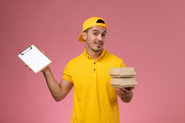 Mensajero masculino de vista frontal en uniforme amarillo sosteniendo pequeños paquetes de alimentos y bloc de notas pensando en el fondo rosa claro.