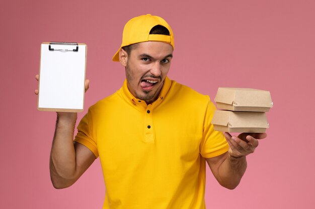 Mensajero masculino de vista frontal en uniforme amarillo sosteniendo pequeños paquetes de alimentos y un pequeño bloc de notas en el fondo rosa.